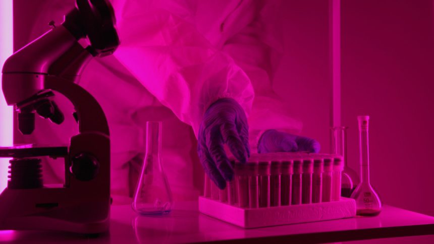 Cientista de jaleco manipula tubos de ensaio diante de um microscópio em um laboratório.