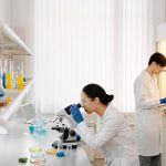 Dois pesquisadores trabalham com microscópios e tubos de ensaio em um laboratório equipado.