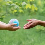Duas mãos trocam um pequeno globo, simbolizando cooperação e união internacional.